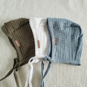 Può includere: Tre cappellini per neonati in verde oliva, bianco e grigio-azzurro, realizzati in un tessuto strutturato. Ogni cappellino ha una piccola etichetta marrone e lacci. I cappellini sono esposti su una superficie chiara e strutturata.