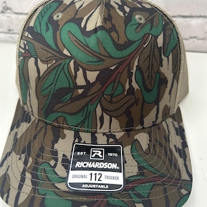 Op de afbeelding: Een camouflage trucker pet met een bladerenpatroon in groen, bruin en tan tinten. De pet heeft een mesh rug en een gebogen klep. De voorkant van de pet heeft een patch met de tekst "RICHARDSON ORIGINAL 112 TRUCKER ADJUSTABLE."