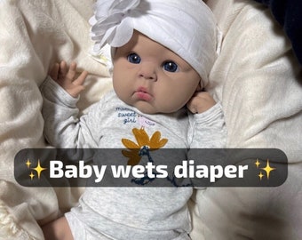 Puede incluir: Una muñeca bebé con un gorro blanco con una flor, un mono gris con el texto "mama sweet girl" y calcetines blancos. La muñeca está tumbada sobre una manta blanca. El texto "Baby wets diaper" está escrito en la imagen.