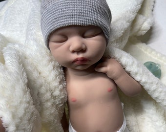 Puede incluir: Un muñeco de bebé durmiendo con un gorro de punto gris y un pañal blanco. El muñeco está acostado sobre una manta blanca y esponjosa.