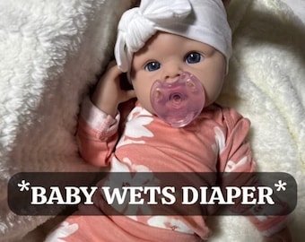 Puede incluir: Una muñeca de bebé de color rosa y blanco con estampado floral que lleva una diadema blanca y un chupete. La muñeca está tumbada sobre una manta blanca y esponjosa. El texto "*BABY WETS DIAPER*" está escrito en la imagen.