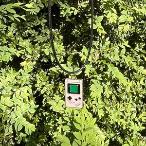 May include: A silver chain necklace with a black cord and a pendant shaped like a vintage handheld video game console. The pendant is white with a green screen.