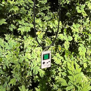 May include: A black cord necklace with a silver chain and a silver pendant shaped like a vintage handheld video game console. The pendant has a green screen and black buttons.