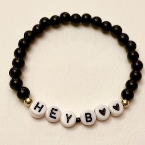 Könnte beinhalten: Schwarzes Perlenarmband mit goldenen Abstandshaltern und weißen Buchstabenperlen, die "HEY B" mit einem Herzsymbol buchstabieren.