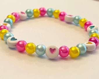 Può includere: Un braccialetto di perline colorate con perline bianche a forma di cuore. Il braccialetto è realizzato con perline rosa, gialle e blu.