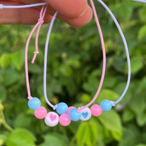 Op de afbeelding: Twee vriendschapsarmbanden met roze en blauwe kralen en kleine hartjesbedels. De armbanden zijn aan elkaar gebonden en worden in een hand gehouden.
