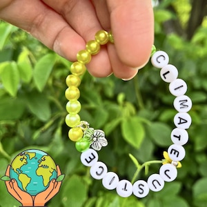 Può includere: Un braccialetto di perline verdi e gialle con le parole "Climate Justice" scritte su perline bianche. Il braccialetto ha un piccolo ciondolo a forma di fiore.
