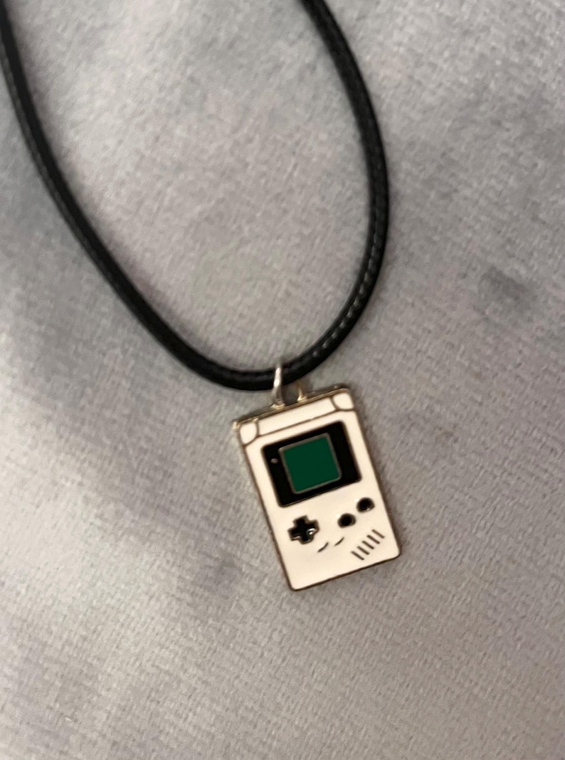 May include: A black cord necklace with a white enamel pendant shaped like a vintage handheld video game console. The pendant has a green screen and gold accents.
