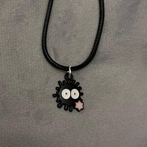 May include: A black necklace featuring a charm shaped like a soot sprite. The charm has white eyes and a pink star. The necklace is on a black cord.