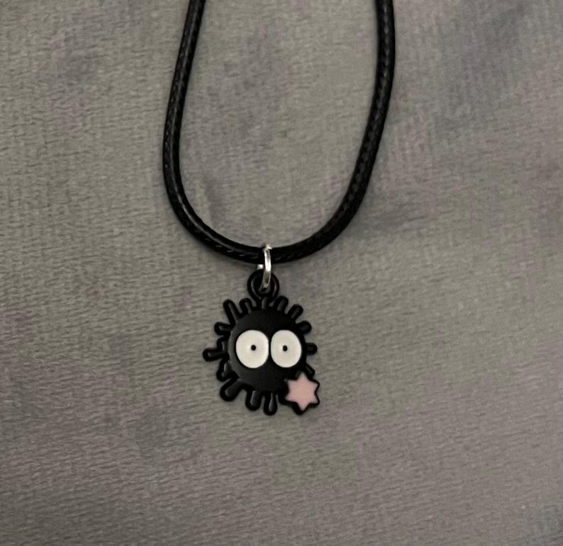 May include: A black necklace featuring a charm shaped like a soot sprite. The charm has white eyes and a pink star. The necklace is on a black cord.