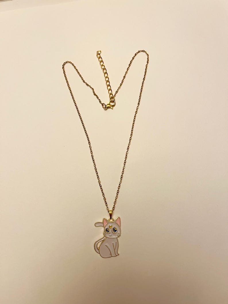 Op de afbeelding: Een gouden ketting met een witte emaille kattenhanger. De kat heeft roze oren en een gouden halve maan op zijn voorhoofd.