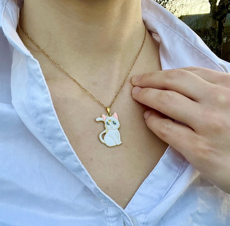 Op de afbeelding: Een gouden ketting met een witte emaille hanger met een cartoonkat met roze oren en blauwe ogen. De kat draagt een gouden kroon.