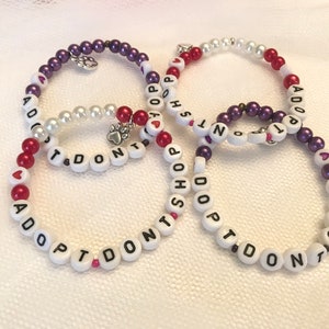May include: Four beaded bracelets with the message "I Don't Shop Adopt" in white beads with red and purple accents. The bracelets have small heart charms.