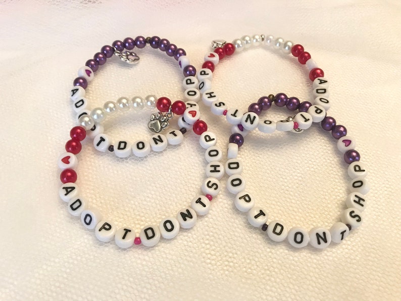 May include: Four beaded bracelets with the message "I Don't Shop Adopt" in white beads with red and purple accents. The bracelets have small heart charms.
