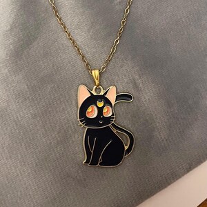 Op de afbeelding: Een gouden ketting met een zwarte kat hanger. De kat heeft roze oren, oranje ogen en een halve maan op zijn voorhoofd.