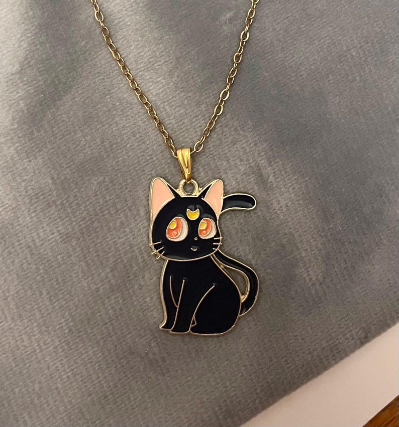 Op de afbeelding: Een gouden ketting met een zwarte kat hanger. De kat heeft roze oren, oranje ogen en een halve maan op zijn voorhoofd.