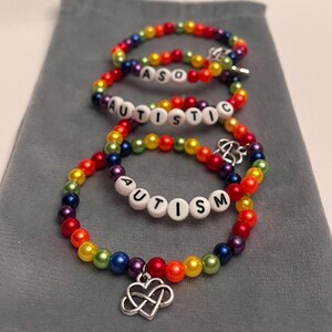 Op de afbeelding: Drie regenboogkleurige armbanden met het woord "AUTISM" gespeld in witte letterkralen. Elke armband heeft een zilveren bedel. De armbanden worden gepresenteerd op een grijze ondergrond.