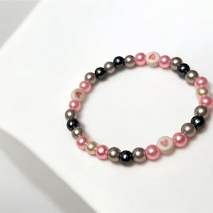 Peut inclure: Un bracelet composé de perles roses, grises et noires. Les perles ont un motif en forme de cœur.