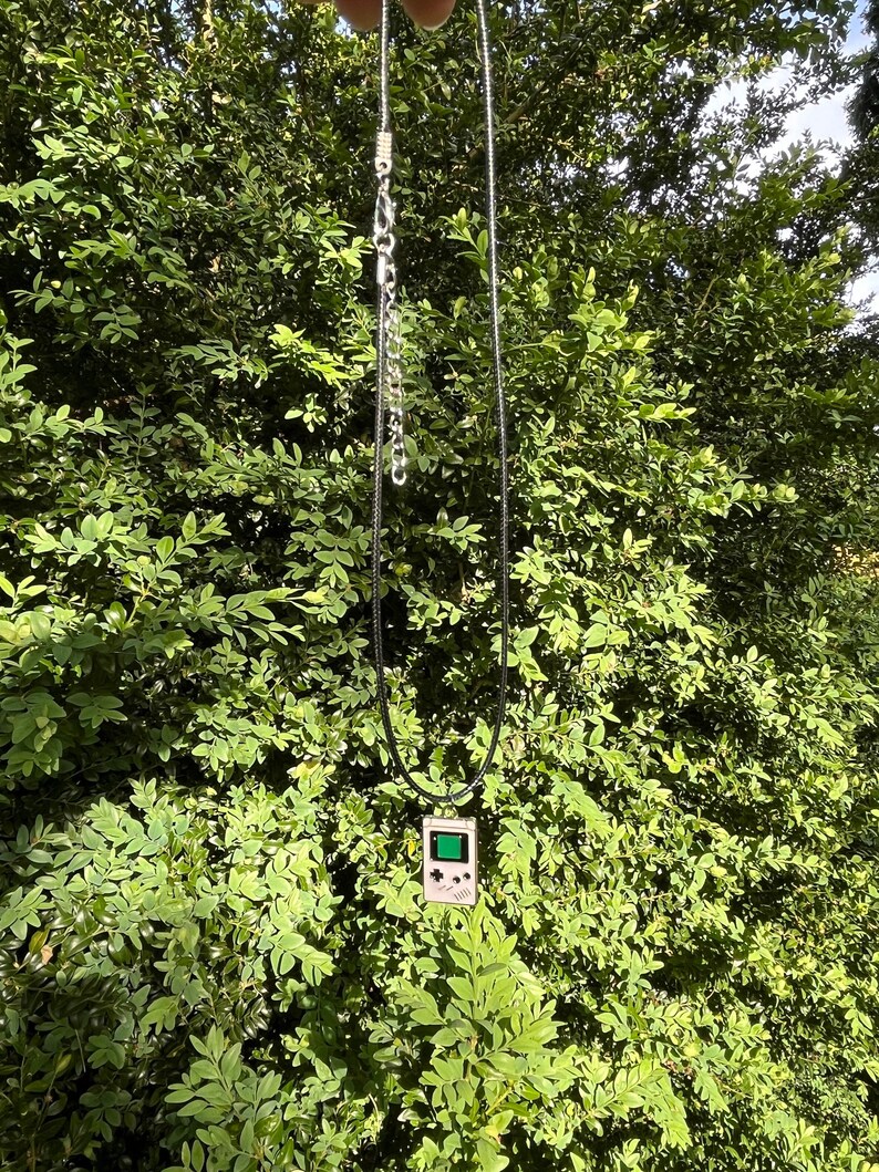 May include: A silver chain necklace with a black cord and a pendant shaped like a vintage handheld video game console. The pendant is white with a green screen.