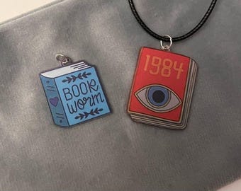 May include: Two pendant necklaces featuring book designs. One is a blue book with the words "BOOK WORM" and a heart. The other is a red book with the number "1984" and an eye design. Both have silver-tone loops.