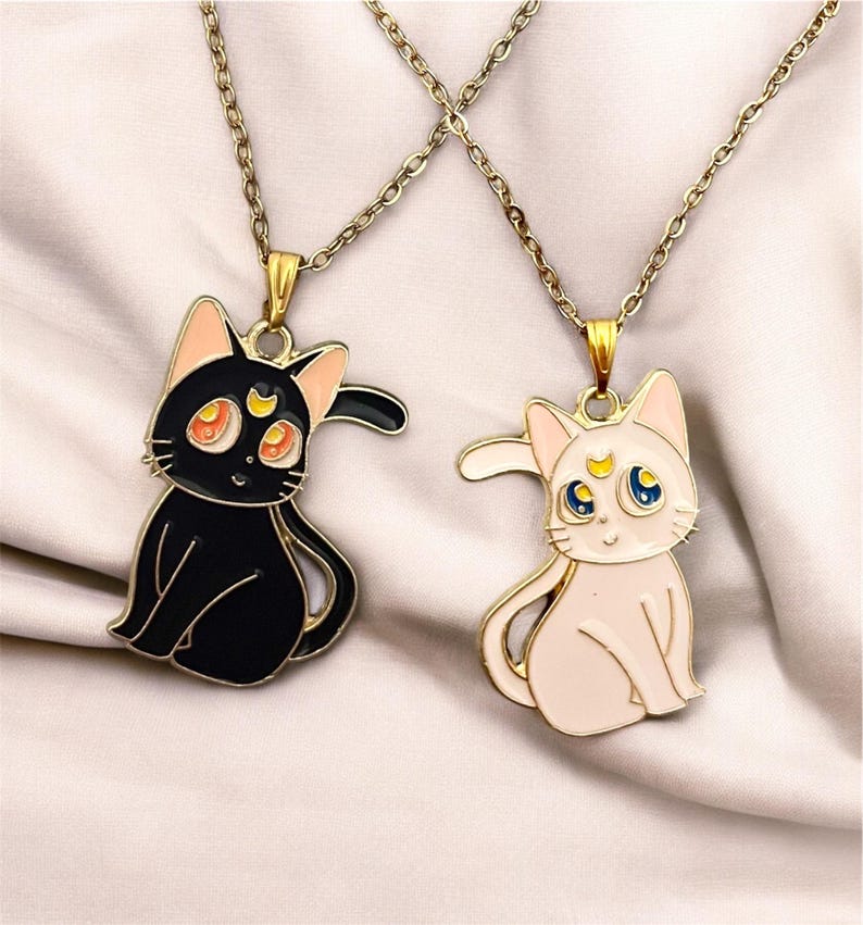 Op de afbeelding: Twee gouden kettingen met zwarte en witte katten hangers. De zwarte kat heeft een halve maan op zijn voorhoofd en oranje ogen. De witte kat heeft een halve maan en een gele ster op zijn voorhoofd en blauwe ogen.