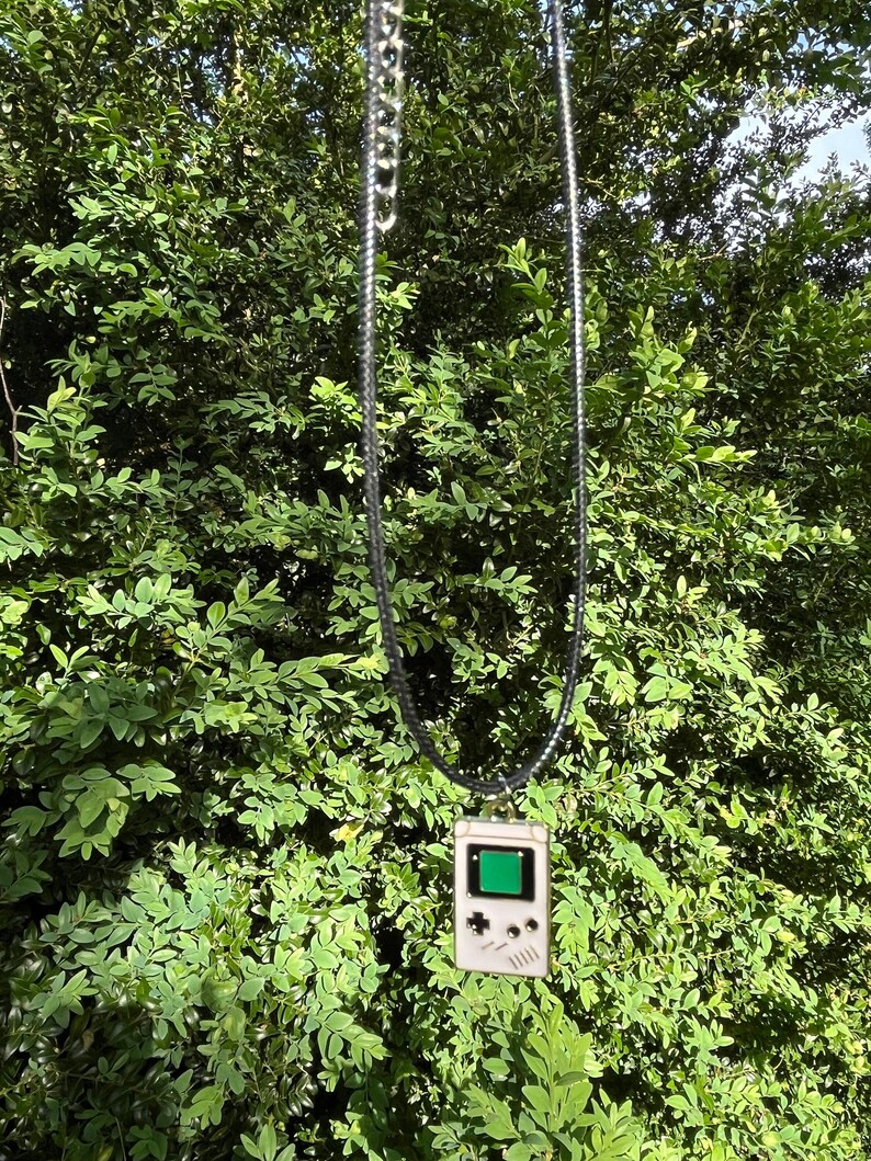 May include: A white and black Game Boy pendant necklace with a green screen. The necklace is hanging from a black cord in front of a green leafy background.