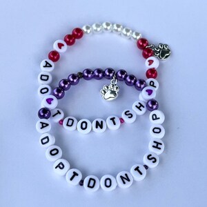May include: Two beaded bracelets with the message "I Don't Shop Adopt" on one and "Adopt Don't Shop" on the other. The bracelets are made with white, purple, and red beads and have silver paw print charms.