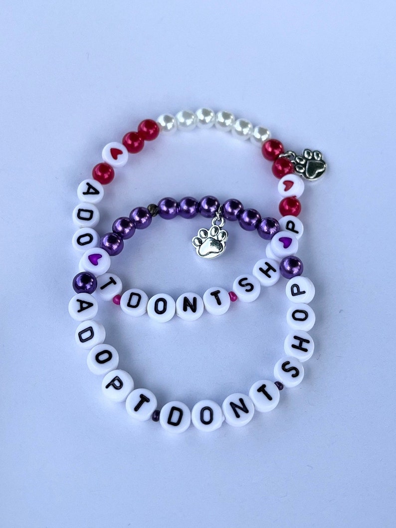 May include: Two beaded bracelets with the message "I Don't Shop Adopt" on one and "Adopt Don't Shop" on the other. The bracelets are made with white, purple, and red beads and have silver paw print charms.