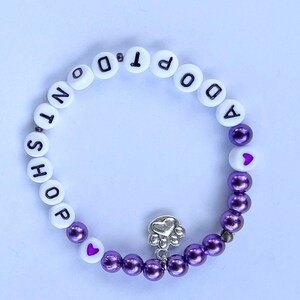 May include: Purple and white beaded bracelet with the words "Adopt Don't Shop" and a silver paw print charm.