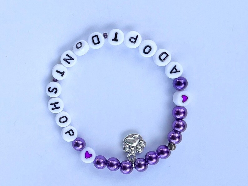 May include: Purple and white beaded bracelet with the words "Adopt Don't Shop" and a silver paw print charm.