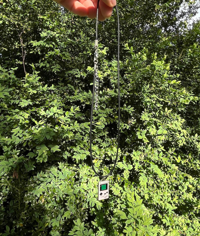 May include: A black cord necklace with a silver chain and a silver pendant shaped like a vintage handheld video game console. The pendant has a green screen and black buttons.