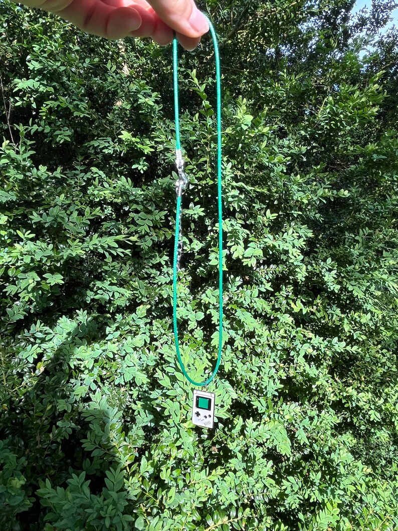 May include: A green cord necklace with a silver clasp and a pendant shaped like a vintage handheld video game console.