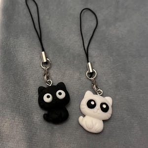 Könnte beinhalten: Zwei katzenförmige Anhänger, einer schwarz und einer weiß, jeweils mit großen, runden Augen. Sie sind an schwarzen Kordeln mit silberfarbenen Metallverschlüssen befestigt. Die Anhänger liegen auf einer grauen Oberfläche.