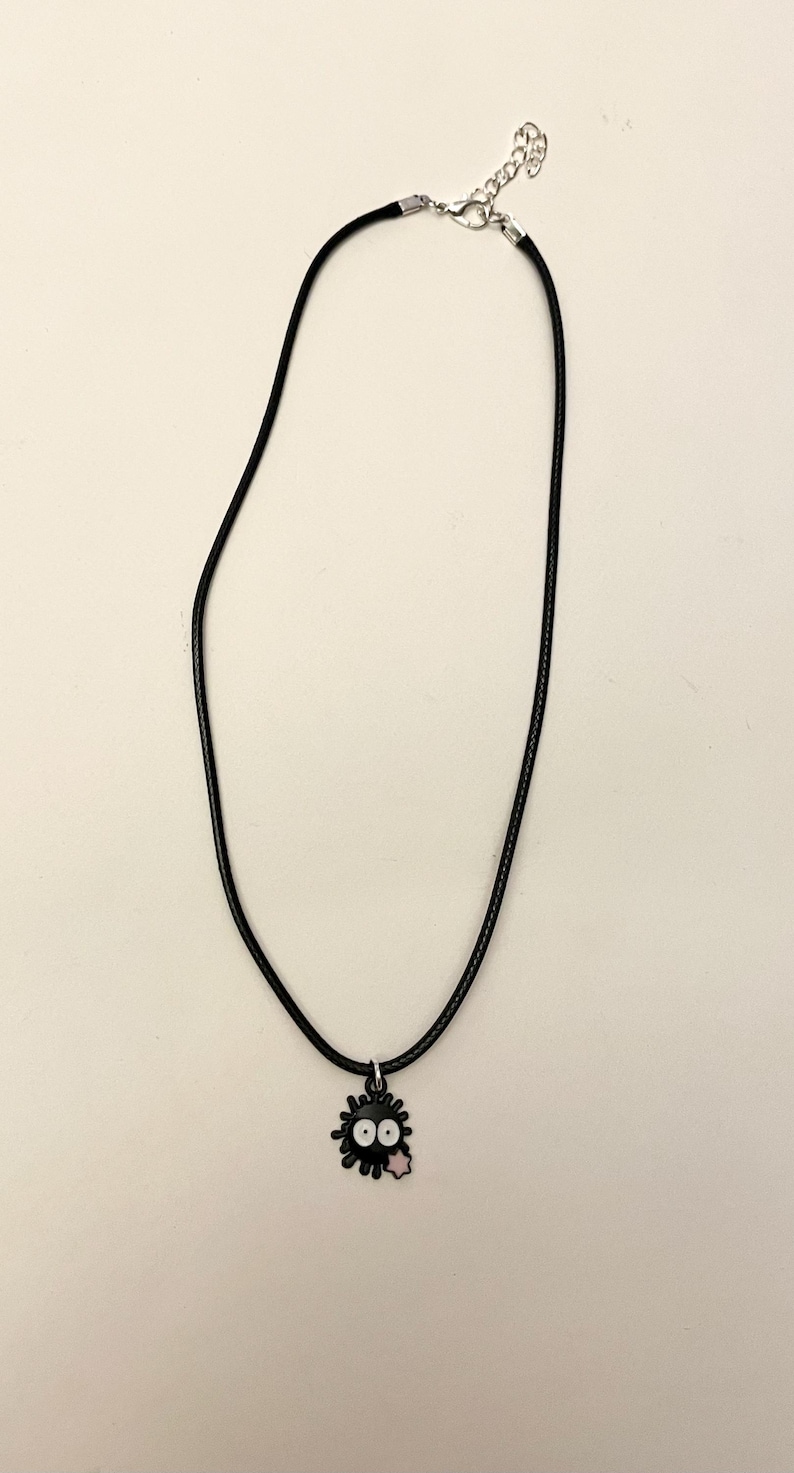 May include: Black cord necklace with a charm featuring a black soot sprite with white eyes and a pink tongue. The necklace has a silver-colored clasp and is displayed against a white background.