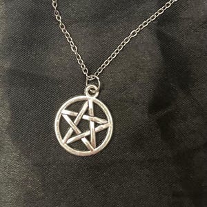 Puede incluir: Collar con colgante plateado con un diseño de pentagrama dentro de un marco circular. El collar tiene una cadena fina y se muestra sobre un fondo oscuro. El colgante es un símbolo a menudo asociado con la Wicca y el paganismo.