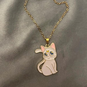 Op de afbeelding: Een gouden ketting met een witte emaille hanger met een cartoonkat met een halve maan en een ster op zijn voorhoofd. De kat heeft blauwe ogen en roze oren.