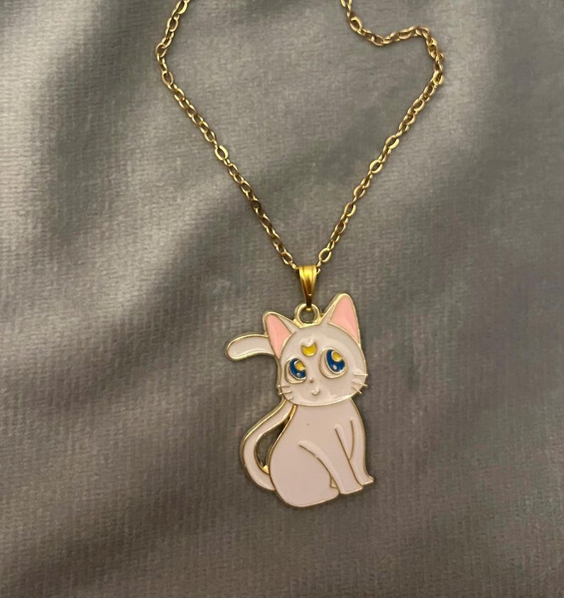 Op de afbeelding: Een gouden ketting met een witte emaille hanger met een cartoonkat met een halve maan en een ster op zijn voorhoofd. De kat heeft blauwe ogen en roze oren.