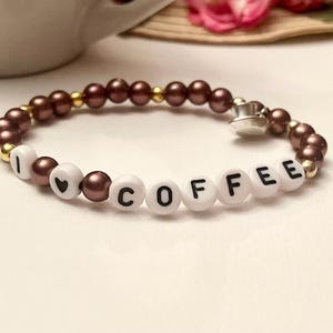 Könnte beinhalten: Ein braunes Perlenarmband mit goldener Akzent und weißen Buchstabenperlen, die "Ich liebe Kaffee" buchstabieren.