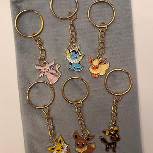 May include: Six gold-colored keychains with colorful enamel charms featuring various cartoon characters. The charms are attached to the key rings with small gold chains. The keychains are resting on a gray velvet pouch.