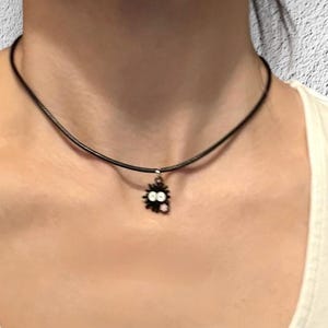 May include: A black cord necklace with a small, black charm featuring a cartoon-like creature with white eyes. The necklace is worn around the neck, against a white shirt.