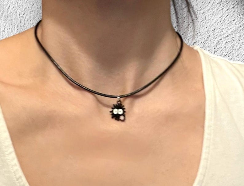 May include: A black cord necklace with a small, black charm featuring a cartoon-like creature with white eyes. The necklace is worn around the neck, against a white shirt.