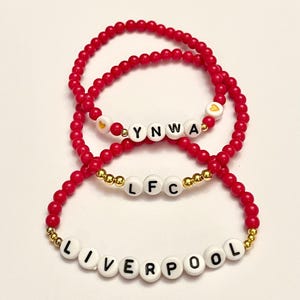 May include: Three red beaded bracelets with gold beads and white letter beads spelling out "LIVERPOOL", "LFC", and "YNWA" with a gold heart bead on each side of "YNWA".