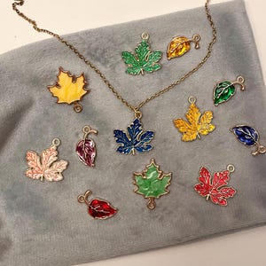 May include: A gold necklace featuring a blue maple leaf pendant, alongside several colourful leaf charms. The charms are in green, yellow, red, and pink, with gold edging, all displayed on a grey surface.