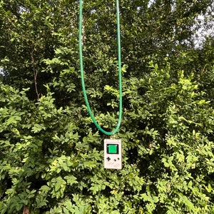 May include: A green cord necklace with a white Game Boy pendant. The pendant has a green screen and black buttons.
