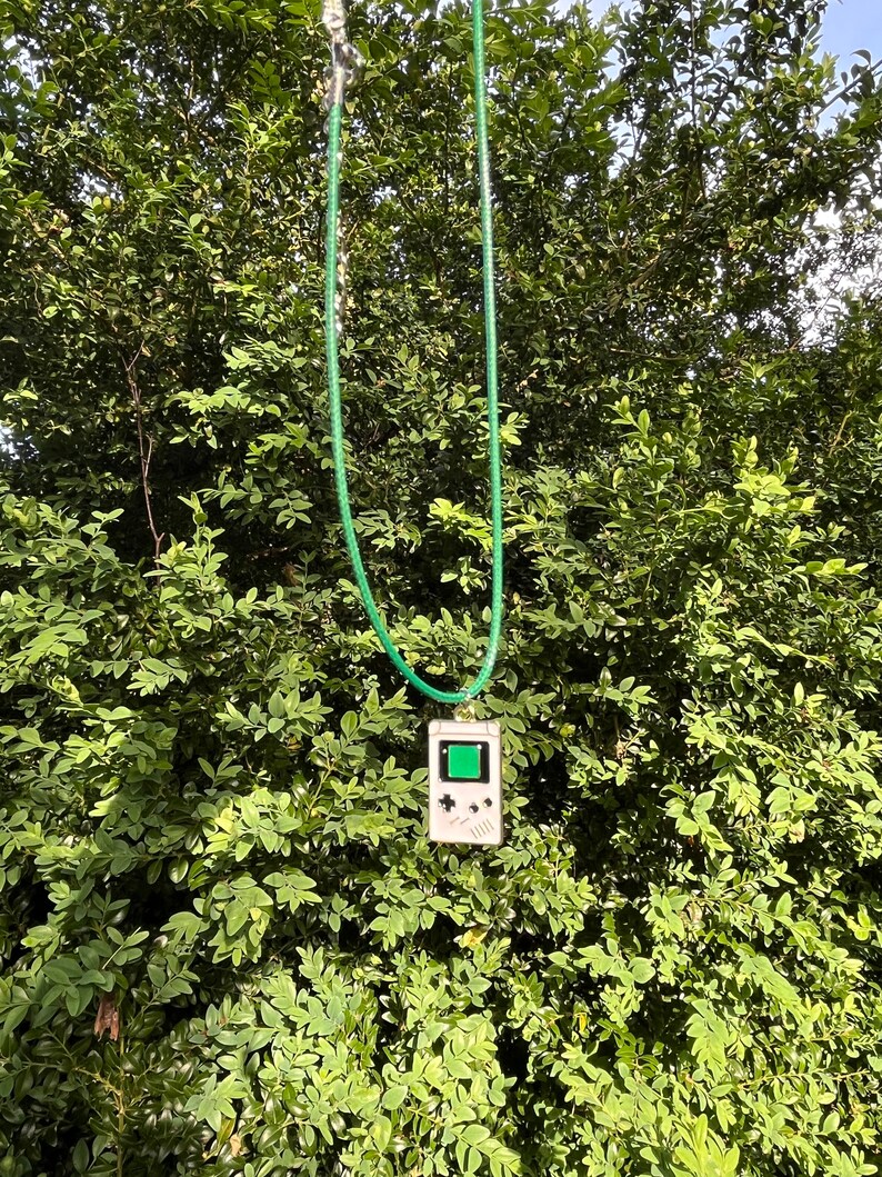 May include: A green cord necklace with a white Game Boy pendant. The pendant has a green screen and black buttons.