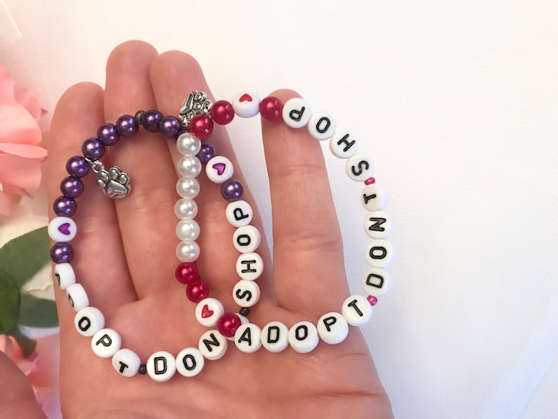 May include: Two beaded bracelets with the words "Don't Shop Adopt" and "Shop Adopt Don't" on them. The bracelets are made with white, red, and purple beads.