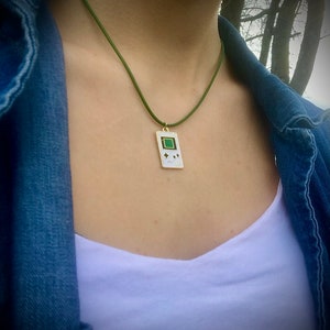 May include: A green cord necklace with a white pendant shaped like a handheld video game console. The pendant has a green screen and black buttons.