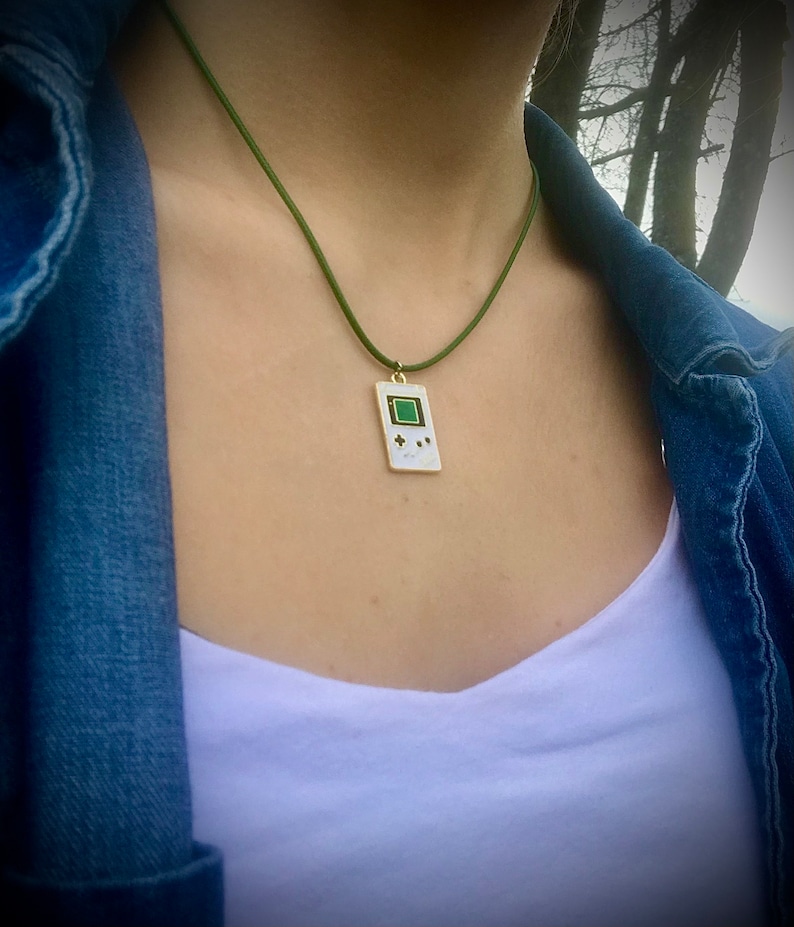 May include: A green cord necklace with a white pendant shaped like a handheld video game console. The pendant has a green screen and black buttons.