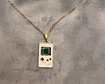 May include: A gold-colored necklace with a pendant shaped like a classic handheld video game console. The pendant is white with black and green details. The necklace is on a gray surface.