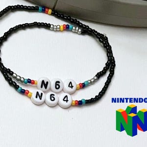 Könnte beinhalten: Eine schwarze Perlenkette mit bunten Perlen und weißen Buchstabenperlen, die "N64" buchstabieren. Die Kette liegt auf einer weißen Oberfläche mit dem Nintendo 64-Logo im Hintergrund.